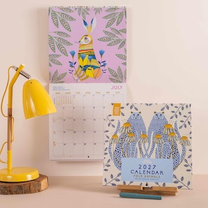 Könnte beinhalten: Ein Wandkalender für 2027 mit dem Titel Folk Animals von Ashley Percival, der eine violette Seite mit einer Hasenillustration und eine weiße Seite mit zwei blauen, gepunkteten Geparden zeigt. Eine gelbe Schreibtischlampe steht auf einem Holzfuß.
