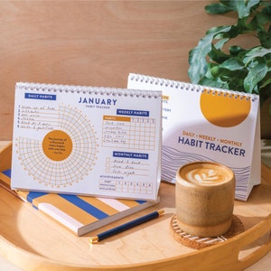 Puede incluir: Un primer plano de un calendario de escritorio con un gráfico de sol amarillo. El calendario se titula "Seguimiento de hábitos diario, semanal y mensual". El calendario está abierto a enero y tiene secciones para hábitos diarios, semanales y mensuales. Una taza de café se encuentra en una bandeja de madera junto al calendario.