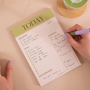 Könnte beinhalten: Ein Tagesplaner-Block mit einer hellgrünen Kopfzeile mit der Aufschrift TODAY. Die Seite enthält Abschnitte für PRIORITIES, TO DO, SCHEDULE und MUST REMEMBER mit handschriftlichen Einträgen. Ein lavendelfarbener und grüner Stift liegt auf dem Papier.