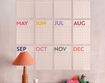 Puede incluir: Doce calendarios mensuales con cuadrículas y nombres de meses en colores vibrantes. Se exhiben en una pared clara sobre una cómoda de madera con una lámpara, una taza y objetos decorativos.