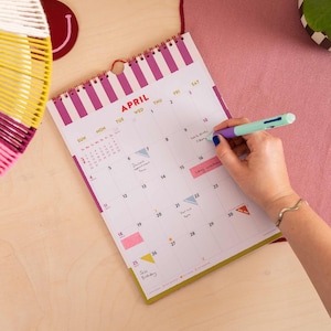 May include: A spiral-bound wall calendar for the month of April featuring vertical magenta and white stripes at the top. The calendar grid contains handwritten notes in various boxes, and a light blue and purple multi-color pen is writing on it.