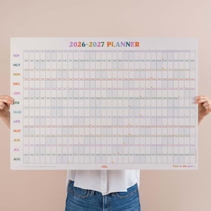 May include: A large white 2026-2027 planner with a grid layout, featuring months and dates. The title "PLANNER" is in rainbow colours. The text "This is the year..." is at the bottom.