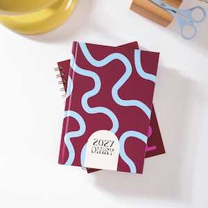 Può includere: Un'agenda bordeaux con un motivo a linee ondulate azzurre e un'etichetta semicircolare bianca con il testo 2027 DIARY. L'agenda è rilegata a spirale e sovrapposta a un'altra agenda identica, con un paio di forbici azzurre nelle vicinanze.