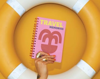 May include: A pink spiral bound notebook with the text "TRAVEL MEMORIES" in yellow letters. The notebook has a red and orange illustration of a palm tree on the cover.