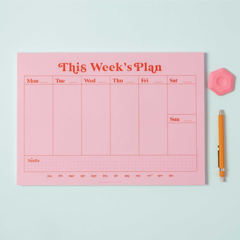 May include: A pink weekly planner with a grid pattern and the text "This Week's Plan" in red. The planner has spaces for each day of the week, Monday through Sunday, and a section for notes. The planner is on a light blue background.