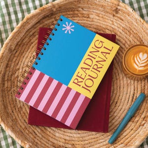 May include: A reading journal with a blue cover and a pink asterisk. The journal has a yellow section with the words "READING JOURNAL" and pink and red stripes. A maroon book is beneath it. A cup of coffee and a teal pen are also in the image.