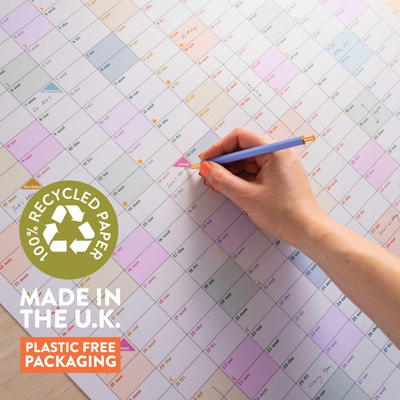 May include: A large, colorful wall calendar with a grid layout, featuring pastel-colored squares for each day. A hand holding a blue pencil is writing on the calendar. The calendar is made from 100% recycled paper and packaged without plastic. Text on the calendar says "Made in the U.K."