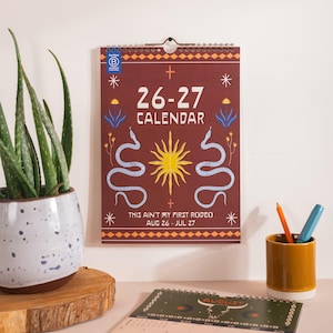 Könnte beinhalten: Ein Wandkalender mit Spiralbindung und dunkelrotem Hintergrund. Er zeigt den Text "26-27 CALENDAR" und "THIS AIN'T MY FIRST RODEO AUG 26 - JUL 27". Das Design umfasst Illustrationen von Schlangen, einer Sonne und floralen Elementen. Ein Blumentopf und ein Stifthalter sind ebenfalls sichtbar.