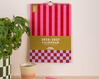 May include: A 2026-2027 wall calendar with a pink and red striped design. The calendar features a gold section with the text "2026-2027 CALENDAR" and a checkered pattern at the bottom. A pink pen and a cup are in the foreground.