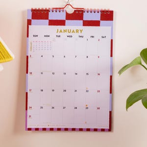 May include: A wall calendar for the month of January featuring a red and light purple checkered border. The calendar includes a grid with days of the week, dates, and a small February preview. It is held together by a red metal spiral binding.