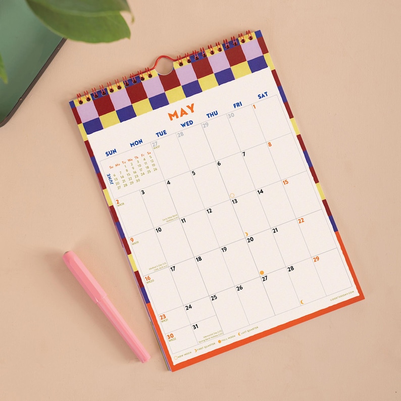 May include: A wall calendar for the month of May featuring a red spiral binding and a checkered border in purple, yellow, and dark blue. The grid displays dates from 1 to 31 with a small June calendar inset. A light pink pen lies next to the calendar.