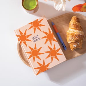 May include: A 2027 diary with a light pink cover featuring a repeating pattern of orange suns. The diary is placed next to a multi-coloured pen and a croissant on a wooden tray, with a small cup of green liquid visible in the upper corner.