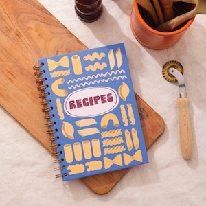 May include: A blue spiral-bound recipe book featuring pasta illustrations and the word "RECIPES" in a white oval. A pasta cutter with a wooden handle is on the right. The book rests on a wooden chopping board.