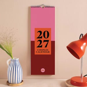 May include: A vertical wall calendar for the year 2027 titled Couples Calendar. The design features a pink top section, a dark red bottom section, and a central orange rectangle with the black text 2027 and the words Couples Calendar below.