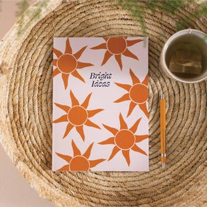 May include: A pink notebook with a repeating pattern of orange suns. The notebook has the text "Bright Ideas" printed on the front.