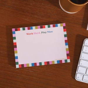 May include: A pink notepad with a colourful striped border. The notepad has the text "Work Hard, Play Nice" printed on the top.