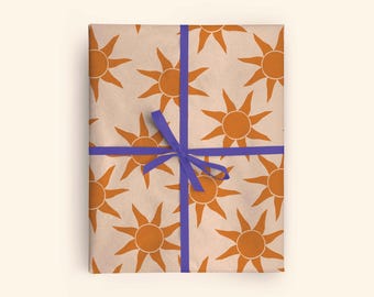 May include: A wrapped gift with a repeating pattern of orange suns on a tan background. The gift is tied with a blue ribbon in a cross pattern and a bow. The overall theme is a cheerful, sunny design.