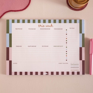 Puede incluir: Un bloc de notas planificador semanal con fondo rosa. El bloc de notas tiene un borde a rayas en tonos marrón, verde y azul. El texto "this week" está en la parte superior, con los días de la semana enumerados debajo. Un bolígrafo rosa está a la derecha.