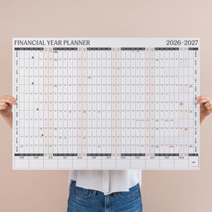 May include: A white Financial Year Planner for 2026-2027, held by a person. The planner features a grid layout with monthly and weekly columns. The text "FINANCIAL YEAR PLANNER" and "2026-2027" are visible.