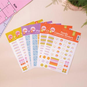May include: A set of colourful family calendar stickers with various designs, including circles, squares, and rectangles. The stickers are printed on recycled paper and feature phrases like "Good Tuesday", "Party", "Trip", and "Weekend".
