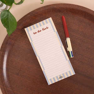 May include: A rectangular to-do list notepad with light blue and cream vertical stripes, featuring the text "to do list" in red script at the top and "GOOD TUESDAY" at the bottom, accompanied by a multi-colour ballpoint pen with a red cap.