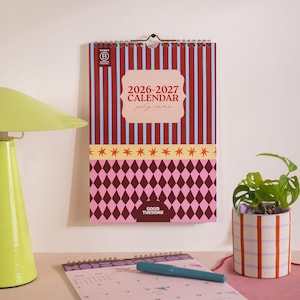 May include: A 2026-2027 wall calendar with a red and blue striped design, a pink diamond pattern, and the text "2026-2027 CALENDAR July-June." The calendar is hanging on a white wall.
