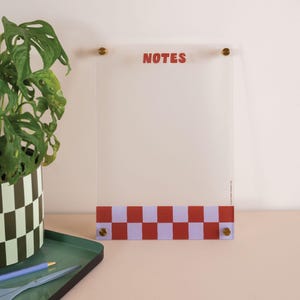 May include: Clear acrylic NOTES memo board with brass hardware. The bottom features a red and lavender checkerboard pattern. A potted plant with a black and green checkered design is in the background.