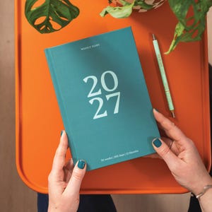 May include: A teal weekly diary with the text 2027 printed in large white font on the front cover, accompanied by the text Weekly Diary and 52 weeks | 365 days | 12 Months. A matching light green pen with text is positioned next to the diary.
