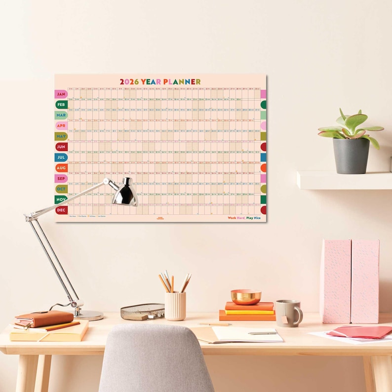 Colorful Year Planner hanging above a stylish desk, showing full year from Jan to Dec 2026 with Moon phases and weekends highlighted.
