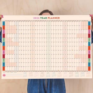 Peut inclure: Un grand planificateur annuel 2026 coloré avec une mise en page en grille. Chaque mois est codé par couleur et comprend un espace pour les notes quotidiennes. Le planificateur est tenu par les mains d'une personne.