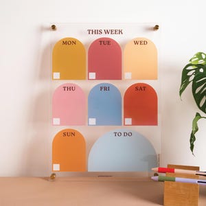 Puede incluir: Planificador semanal de acrílico transparente con secciones arqueadas de colores para cada día de la semana, de Lun a Dom, y una sección "To Do". Montado con herrajes dorados e incluye bolígrafos coloridos.