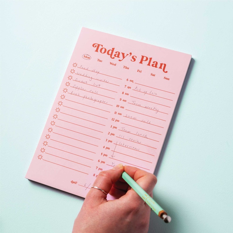 May include: A pink notepad with a red title "Today's Plan" and a daily planner layout. The notepad has a list of tasks written in black ink, including "wedding invites", "pick up Liam", and "team meeting".