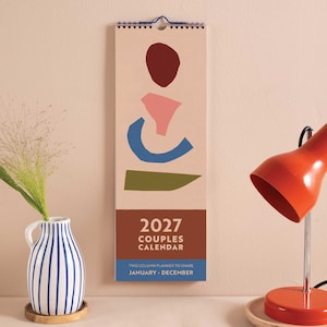 May include: A vertical wall calendar for 2027 titled Couples Calendar. The cover features abstract shapes in dark red, pink, blue, and olive green on a light background. The bottom section includes the text 2027 Couples Calendar and Two Column Planner to Share January - December.