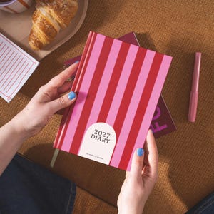 Può includere: Un'agenda con copertina rigida caratterizzata da un motivo a righe verticali che alterna tonalità di rosso e rosa. Un'etichetta bianca ad arco sulla copertina riporta il testo 2027 DIARY e 52 weeks | 12 months in carattere nero.