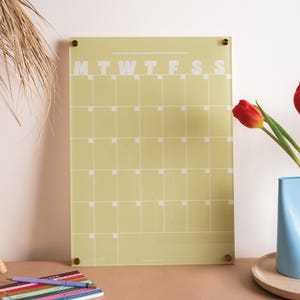 Puede incluir: Un calendario rectangular amarillo pálido con una cuadrícula y los días de la semana impresos en blanco. El calendario está montado sobre una superficie transparente con fijaciones doradas. Un bloc de notas y bolígrafos son visibles en primer plano. Ideal para la planificación semanal.