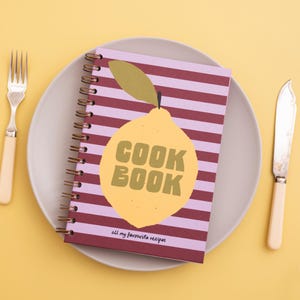 Può includere: Un libro di cucina a spirale con una grafica di limone e la scritta "COOK BOOK" sulla copertina, appoggiato su un piatto con forchetta e coltello. Il libro ha un motivo a righe nei toni del viola e del rosso.