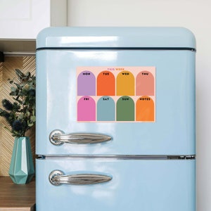 Könnte beinhalten: Ein farbenfroher Wochenplaner-Magnet mit rosa Hintergrund und gewölbten Abschnitten für jeden Wochentag, beschriftet mit Mo, Di, Mi, Do, Fr, Sa, So und Notizen. Die Abschnitte sind in Lila, Orange, Gelb, Pink, Türkis, Grün und Orange gehalten. Der Text "DIESE WOCHE" steht oben.