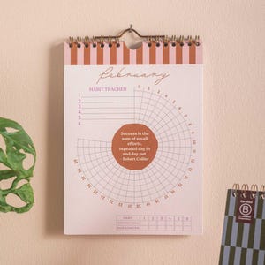 May include: A habit tracker calendar for February, featuring a spiral binding and a striped design in pink and brown. The calendar includes a circular habit tracking chart with numbers and a quote by Robert Collier. The bottom has a section for tracking habits.