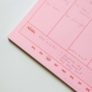 May include: A pink weekly planner with a grid pattern and red lines. The planner has a section for notes and a section for weekly priorities. The months of January through August are listed at the bottom of the planner. The text "Notes" is written in red at the top of the planner. The text "Week priorities" is written in black at the top of the planner. The text "# return books" is written in black at the bottom of the planner. The text "Deadline day" is written in black at the bottom of the planner. The text "Return books" is written in black at the bottom of the planner. The text "Buy bday card" is written in black at the bottom of the planner.