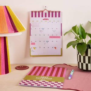 May include: A wall calendar for April 2027 with magenta and white stripes, hanging above a matching desk calendar featuring a checkered pattern. A lamp with a multi-colored yarn shade, a brown smiley face coaster, and a pen are also visible.