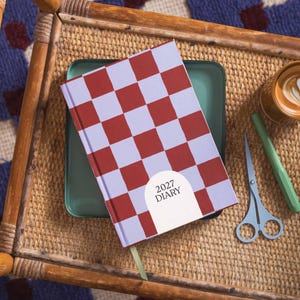 May include: A rectangular 2027 diary featuring a red and light purple checkered pattern. The cover includes a white arch-shaped label with the text 2027 DIARY printed in black. A light green ribbon bookmark extends from the bottom of the diary.