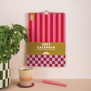 May include: A 2027 wall calendar with a red and pink vertical striped top section, a gold middle band featuring the text 2027 CALENDAR 12 MONTHS JANUARY-DECEMBER GOOD TUESDAY, and a bottom section with a red and light purple checkered pattern.