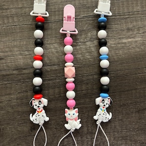 Peut inclure: Trois anneaux de dentition en silicone pour b&eacute;b&eacute; avec clips. Les anneaux de dentition sont noirs et blancs avec des accents rouges, roses et bleus. Un anneau de dentition repr&eacute;sente un dalmatien, un autre un chat et le dernier un dalmatien avec un chapeau bleu.
