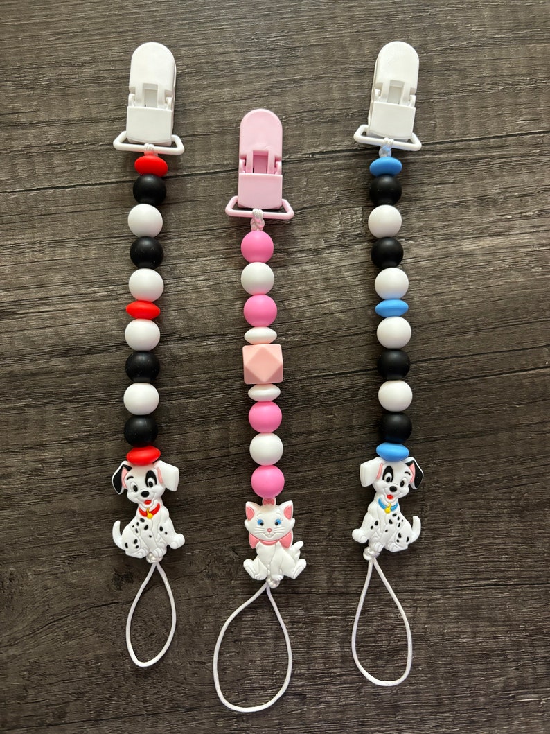 Peut inclure: Trois anneaux de dentition en silicone pour b&eacute;b&eacute; avec clips. Les anneaux de dentition sont noirs et blancs avec des accents rouges, roses et bleus. Un anneau de dentition repr&eacute;sente un dalmatien, un autre un chat et le dernier un dalmatien avec un chapeau bleu.