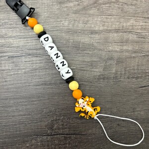 May include: A white silicone baby teether with the name "DANNY" spelled out in black letters. The teether has orange and yellow beads and a black clip. A small orange and black tiger character is attached to the teether.