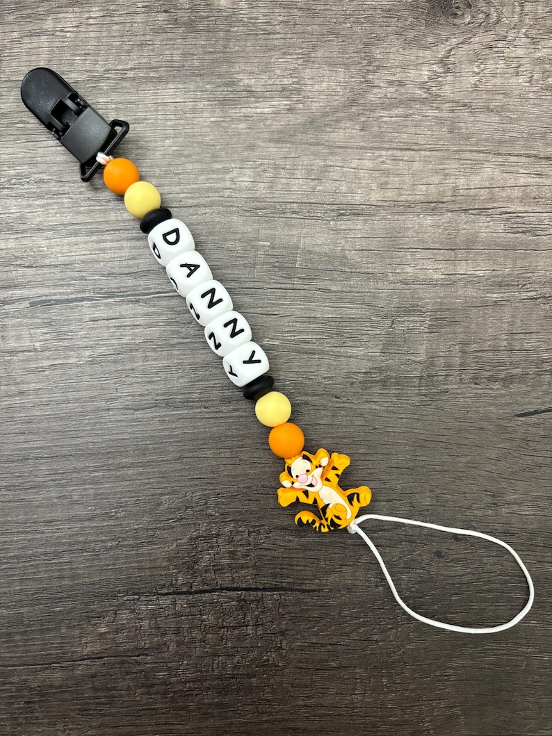 May include: A white silicone baby teether with the name "DANNY" spelled out in black letters. The teether has orange and yellow beads and a black clip. A small orange and black tiger character is attached to the teether.