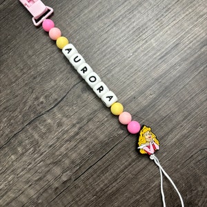 May include: A pink and yellow baby teether with a white clip and a white bead with the name "AURORA" spelled out. The teether also has a pink and yellow silicone charm of Princess Aurora from Sleeping Beauty.