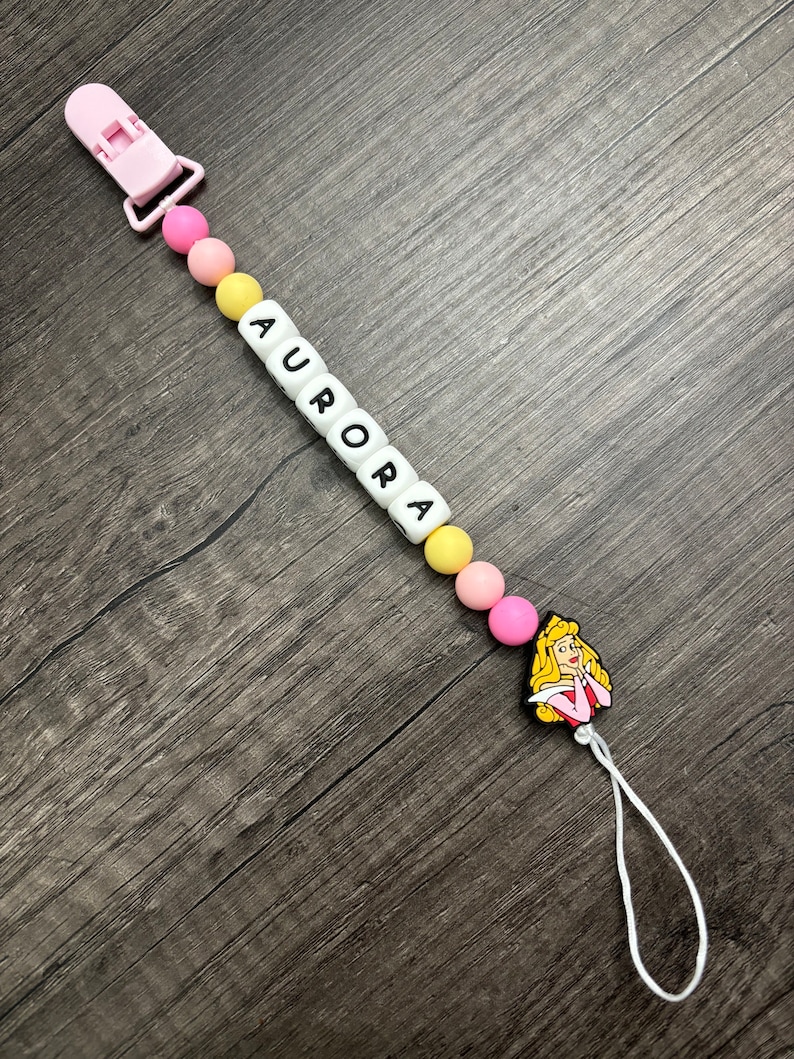 May include: A pink and yellow baby teether with a white clip and a white bead with the name "AURORA" spelled out. The teether also has a pink and yellow silicone charm of Princess Aurora from Sleeping Beauty.
