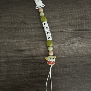 May include: A green and white silicone baby teether with a white clip, a string, and a green and brown character charm. The teether has the name "LUCA" spelled out in black letters.
