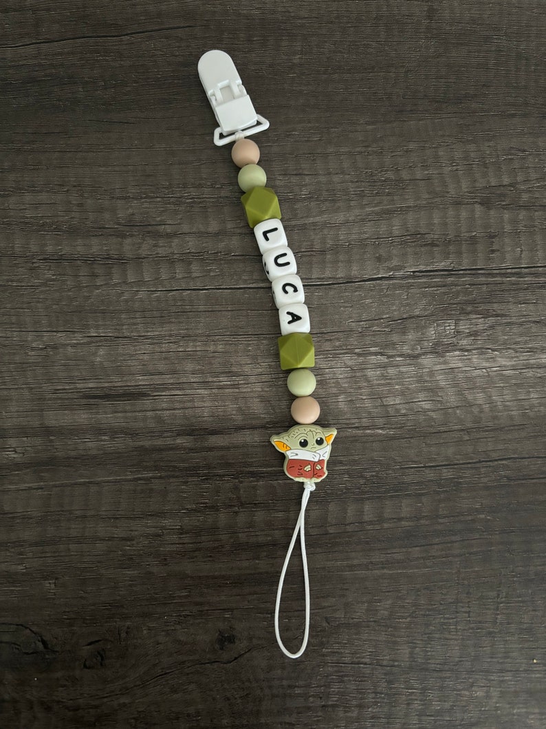 May include: A green and white silicone baby teether with a white clip, a string, and a green and brown character charm. The teether has the name "LUCA" spelled out in black letters.
